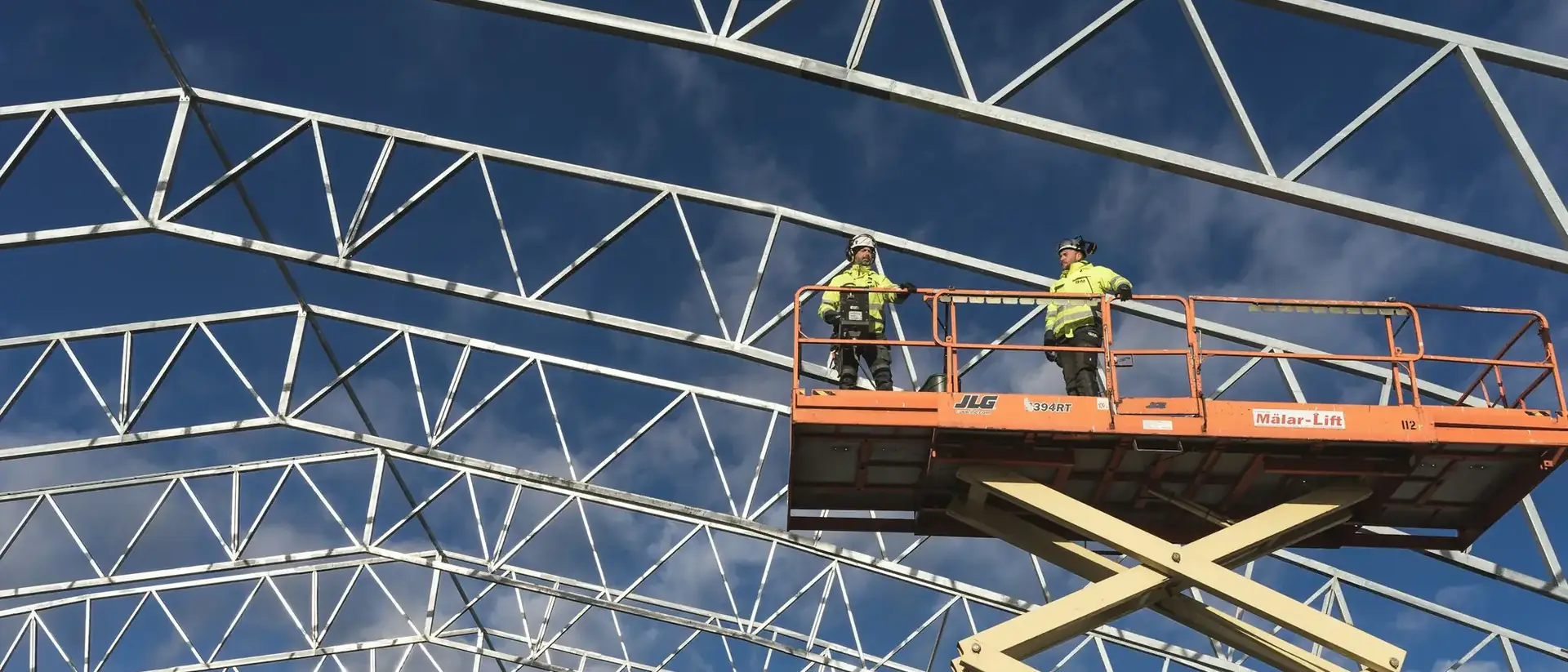 Steel roof trusses being erected by two workers on a scissor lift