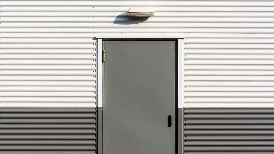 Grey steel door set into a white profiled insulated panel wall with a light fitting above.