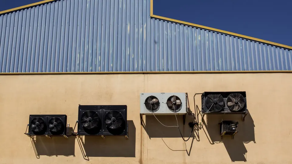 Row of outdoor condensing units with twin fans mounted on a warehouse wall below profiled metal cladding.